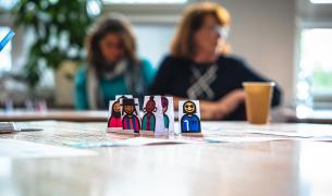 In the foreground are handrawn paper figures of diverse humans on a table while two adult women are visible out of focus in the background seated at another table