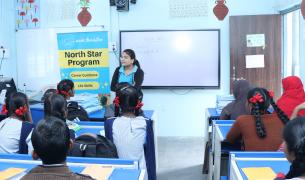 A young woman stands in front of a classroom of school children next to a banner for Asude Foundation's North Star Program