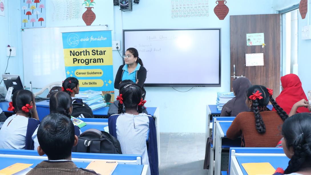 A young woman stands in front of a classroom of school children next to a banner for Asude Foundation's North Star Program