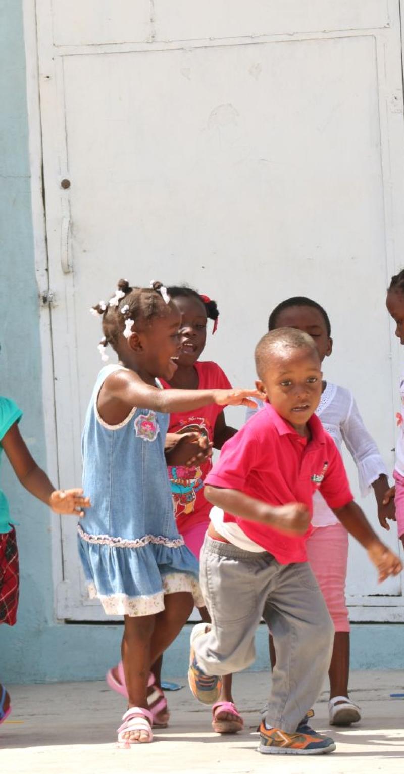 An action shot of several young joyful Black children holding hands in front of a pale blue wall