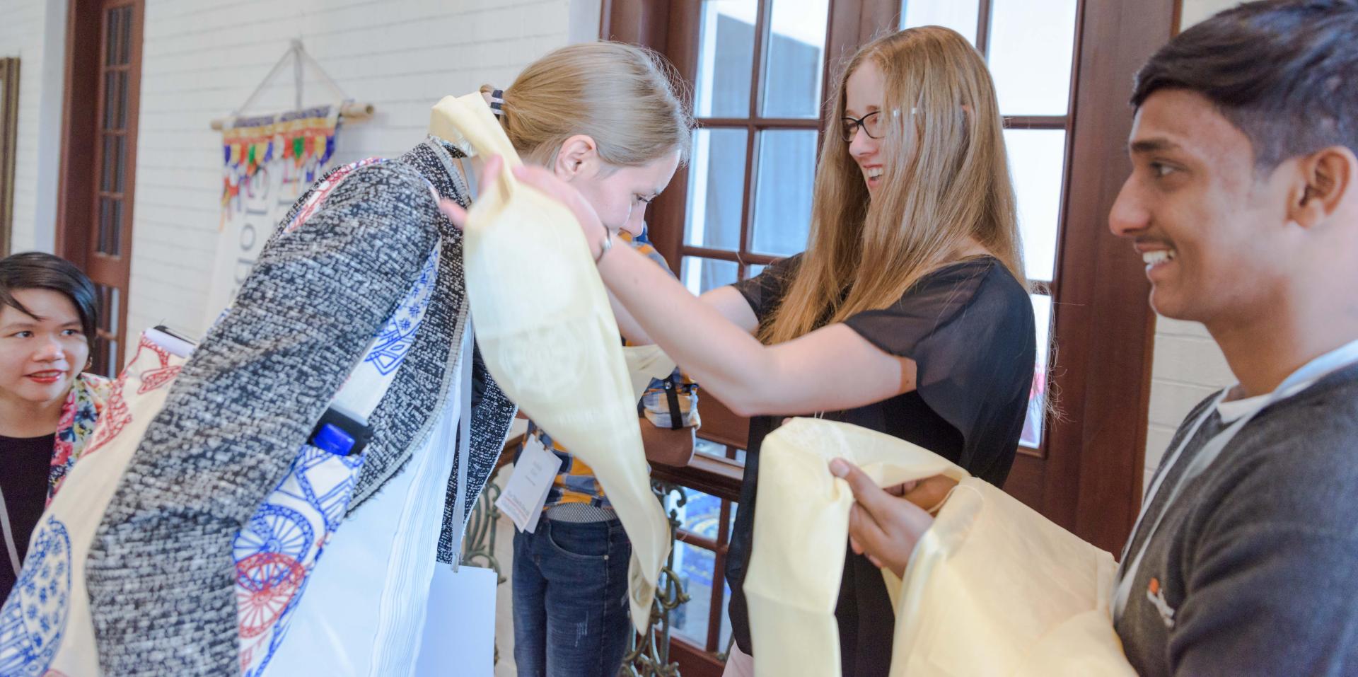 A teenage girl with long red hair places a scarf around the neck of a woman with blond hair. A teenage boy with brown skin smiles as he holds another scarf