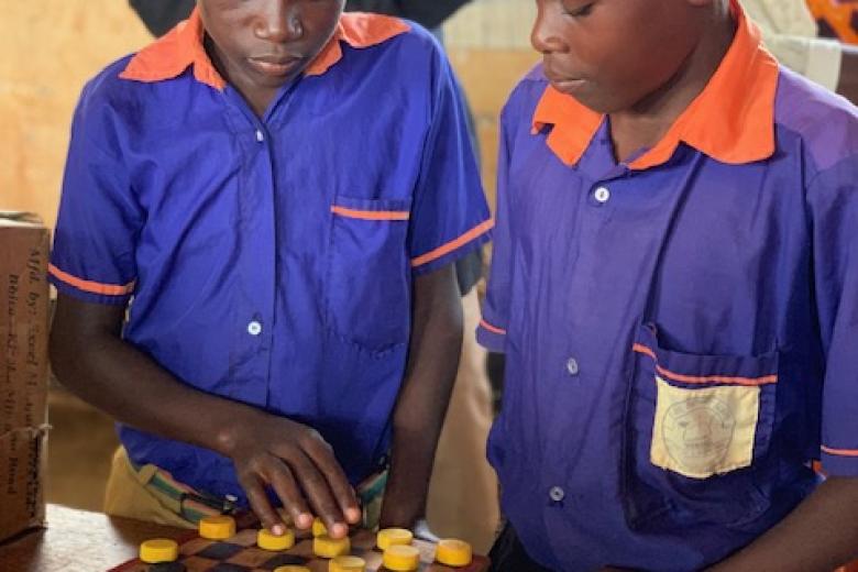 Two young Black boys in blue shirts with orange collars stand in front of a project involving a checker board, one of them touching one of the pieces