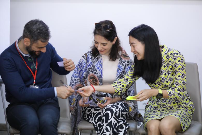 A man in a blue sweater and jeans sits next to two women with long brown hair and they are exchanging sticky notes