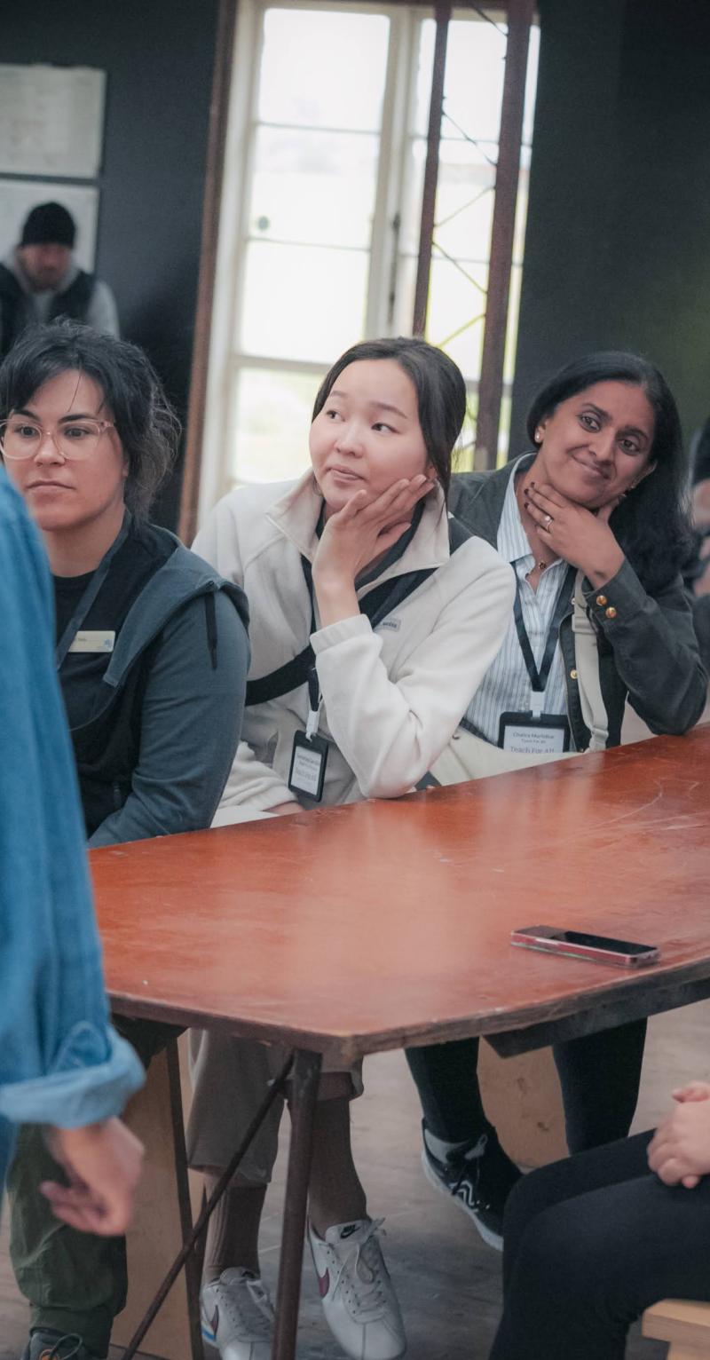 A diverse group of adults sits at a long table looking at woman standing at the end wearing a blue button down and a baseball cap