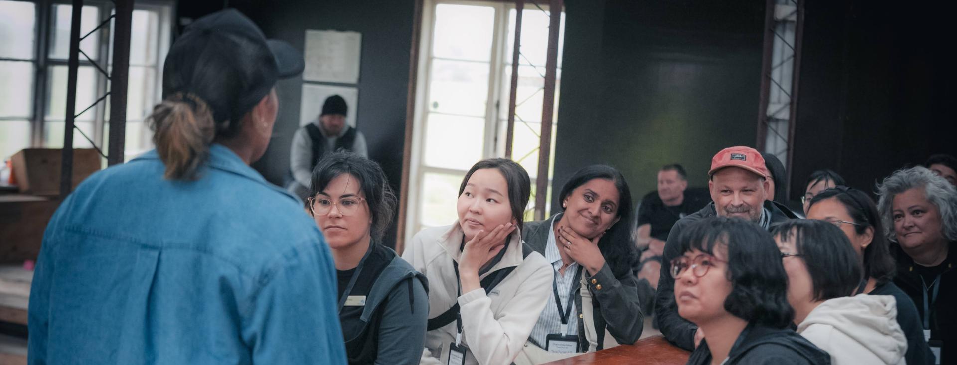 A diverse group of adults sits at a long table looking at woman standing at the end wearing a blue button down and a baseball cap