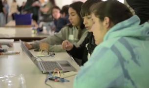 Three teens look at a laptop computer at a table, the girl closest to the camera wears a tie-dyed green sweatshirt.