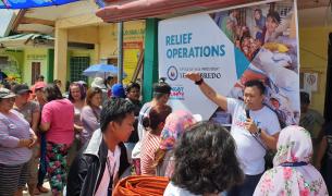 A group of people listen to man holding a microphone speaking in front of a large banner saying "RELIEF OPERATIONS"