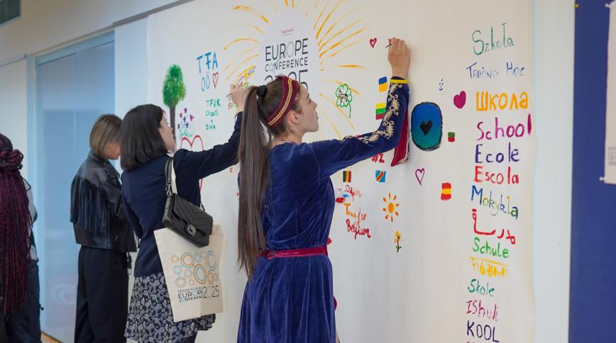 A young woman with a ponytail writes in marker on a wall that says Europe Conference and is covered in brightly colored writing and drawing