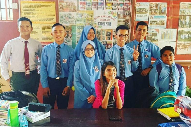 An Asian woman in a pink shirt smiles while seated at table surrounded by teenage students in blue uniforms
