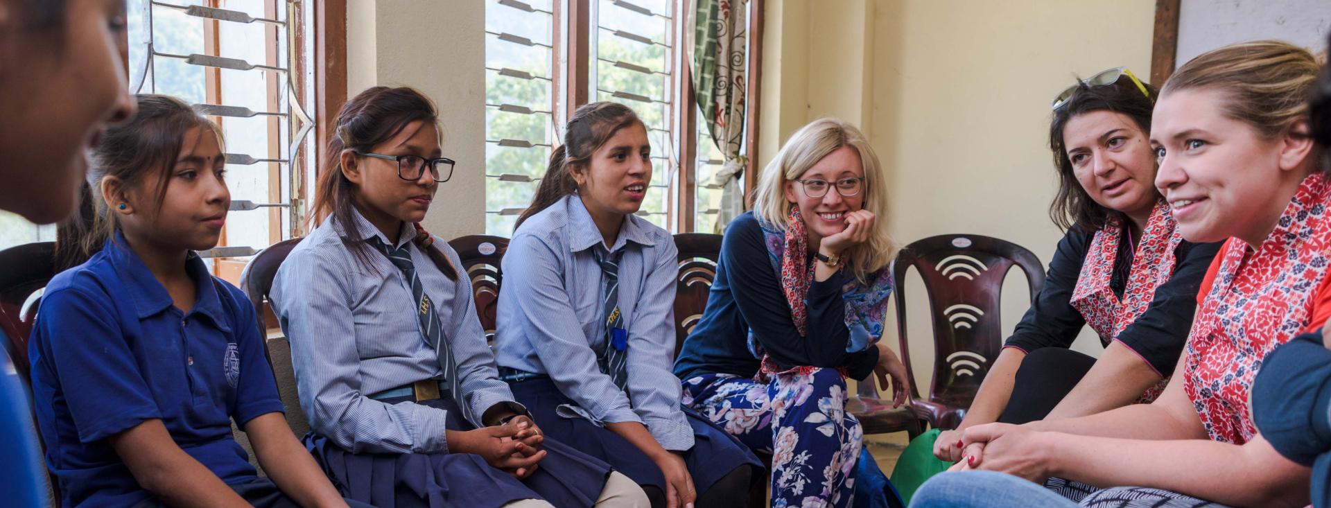 A group of four Asian girls and three European women are seated in a half-circle talking to each other