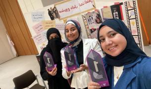 Three women in hijabs pose holding first place trophies in front of a poster with Arabic text and photos on it