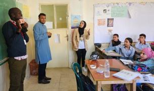 A Black man stands in front of a classroom with his back to the board, holding something up and speaking to students seated at desks
