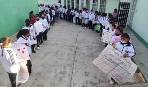 Teenage girls in white shirts holding big posters line two sides of an outdoor walkway between a light green wall and a white wall with windows