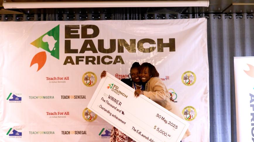 Two Black women hug while holding a giant check in front of a backdrop that says EdLaunch Africa