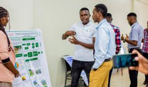 An African man stands in front of an academic poster explaining it to another young man while serveral other people surround them