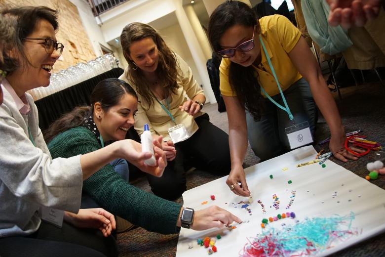 Four women create an art project together with glue, paper, and small colorful objects while seated on the floor