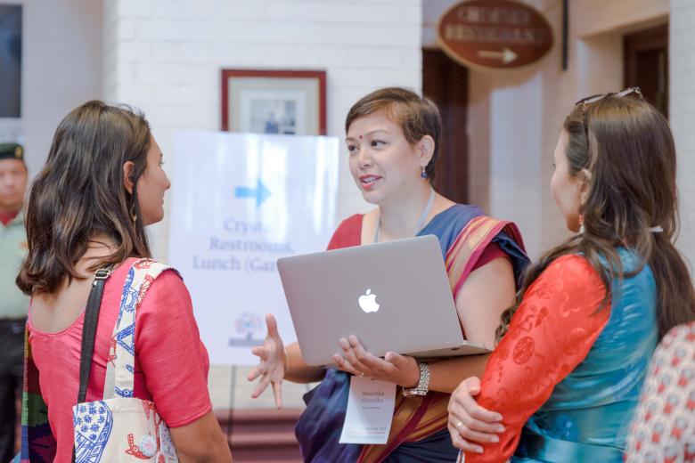 A South Asian woman with short hair holds a laptop computer and speaks to two other women