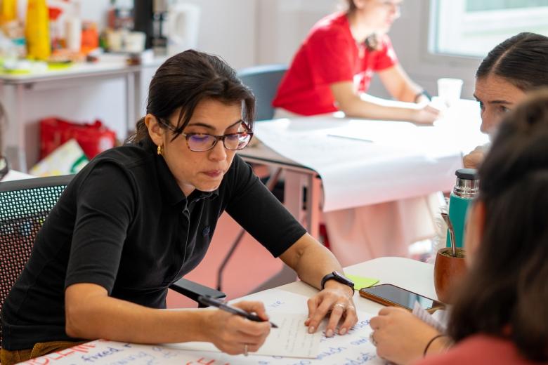 A woman with brown hair and glasses writes on a large piece of paper on a table and we see the backs of two other womens heads