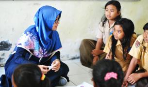 A teacher in a blue hijab sits on the floor talking to young students in their school uniforms