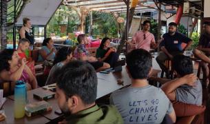 A diverse group of young men and women sit at a table looking towards two men standing at the end of the table speaking