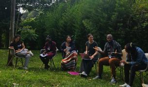 A group of six adults sit in a row on chairs outside on a green grass field in front of some trees