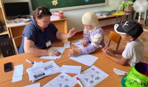 A young female teacher in a blue top shows worksheets to two young students at a table, one wearing a headscarf and the other a baseball cap