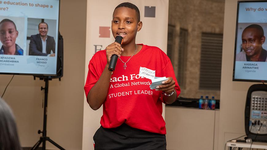 A young african student speaks into a handheld microphone, she is wearing a red shirt that says "Teach For All student leader"