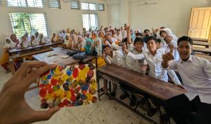 Classroom full of students smiling and posing in the background while in the foreground a hand holds up a small painting of colorful dots