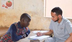 A black girl in a plaid school uniform and a young Black man in a grey Teach For Niger polo shirt look at a notebook together