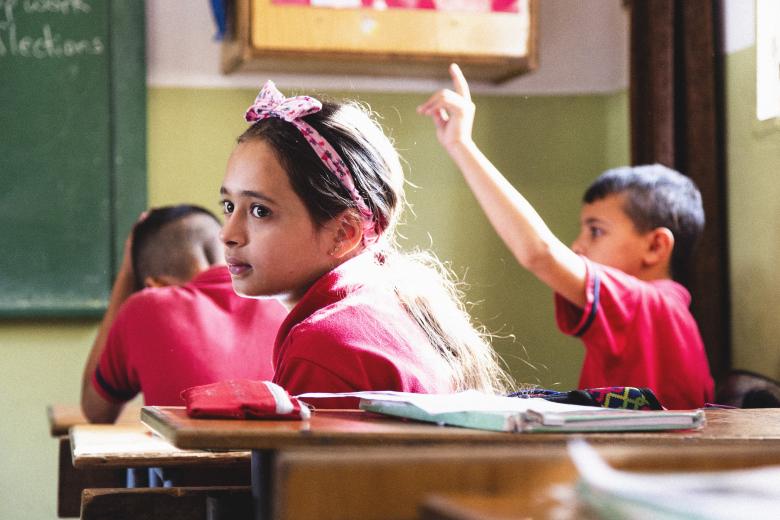 A young girl wearing a headband seated at a desk in a classroom looks off to the left. Two boys sit in the next row, one with his hand raised