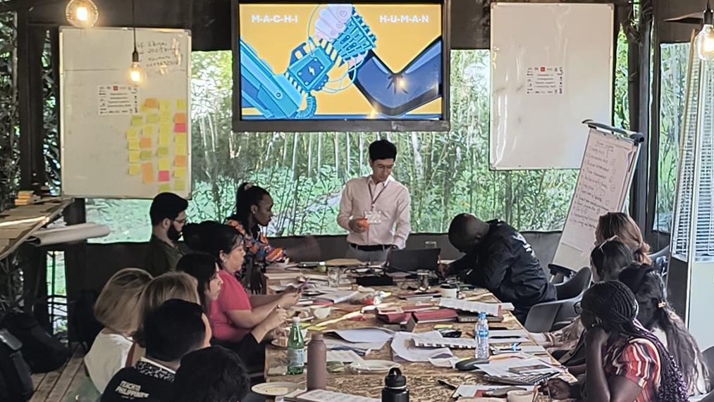 A man stands at the head of a long table with people sitting on either side, there is a screen behind him with a colorful presentation and white board and flipcharts with writing on either side