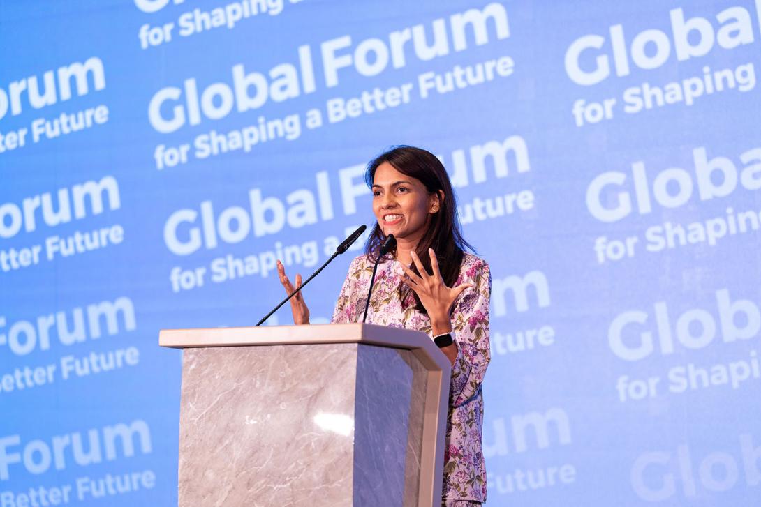 A young woman with long brown hair and tan skin stands at a podium in front of a background that says Global Forum for Shaping a Better Future
