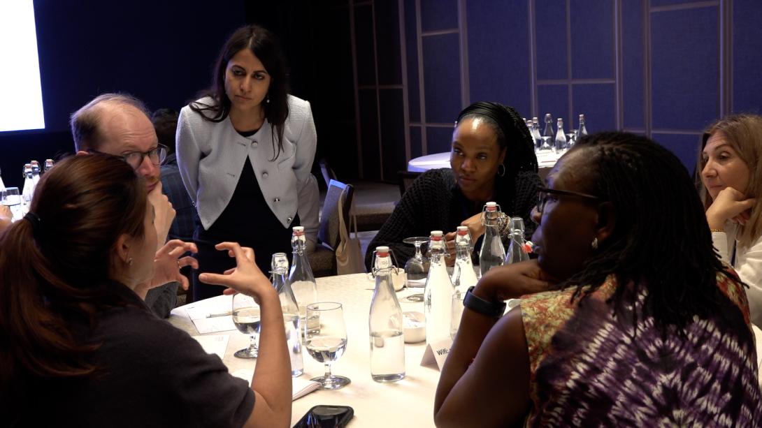 A woman with long brown hair wearing a white blazer leans over a table of diverse adults who appear to be in discussion
