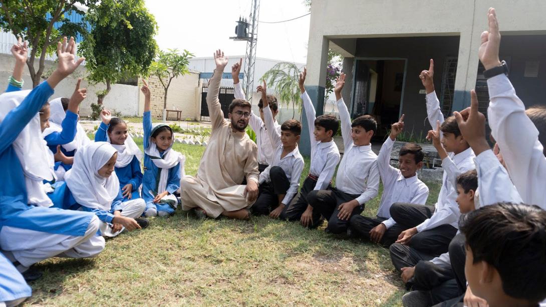 School children in uniform sit in a circle outside on grass with their teacher in the middle, all of them are raising one hands