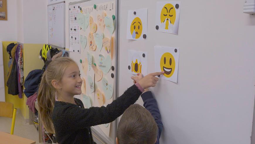 A young girl and boy stand in front of a white board pointing at printed out pictures of emojis representing different feelings