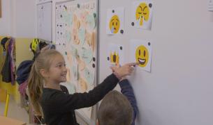 A young girl and boy stand in front of a white board pointing at printed out pictures of emojis representing different feelings