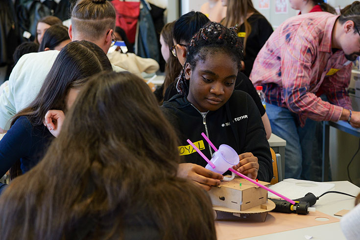 A Black girl sits at a table with two other girls, creating something out of a plastic cup and pipe cleaners