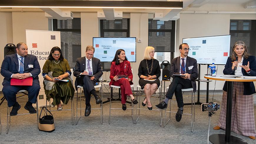 Six people in business clothes seated on high stools with one woman standing and speaking into a mic at the right hand side