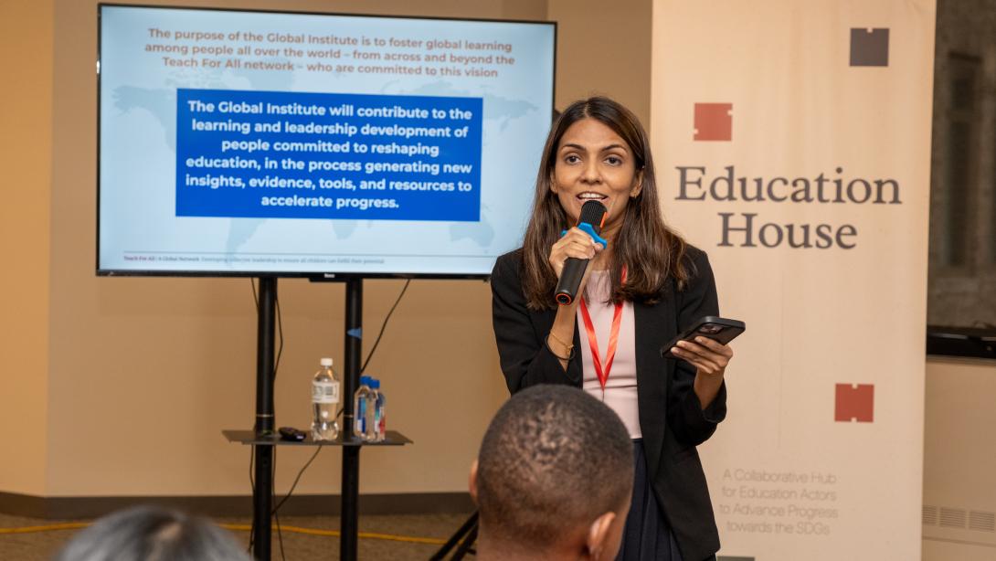 An Asian woman in a blazer speaks into a handheld microphone standing in front of a banner that says Education House and a screen displaying an out-of-focus slide with text on it
