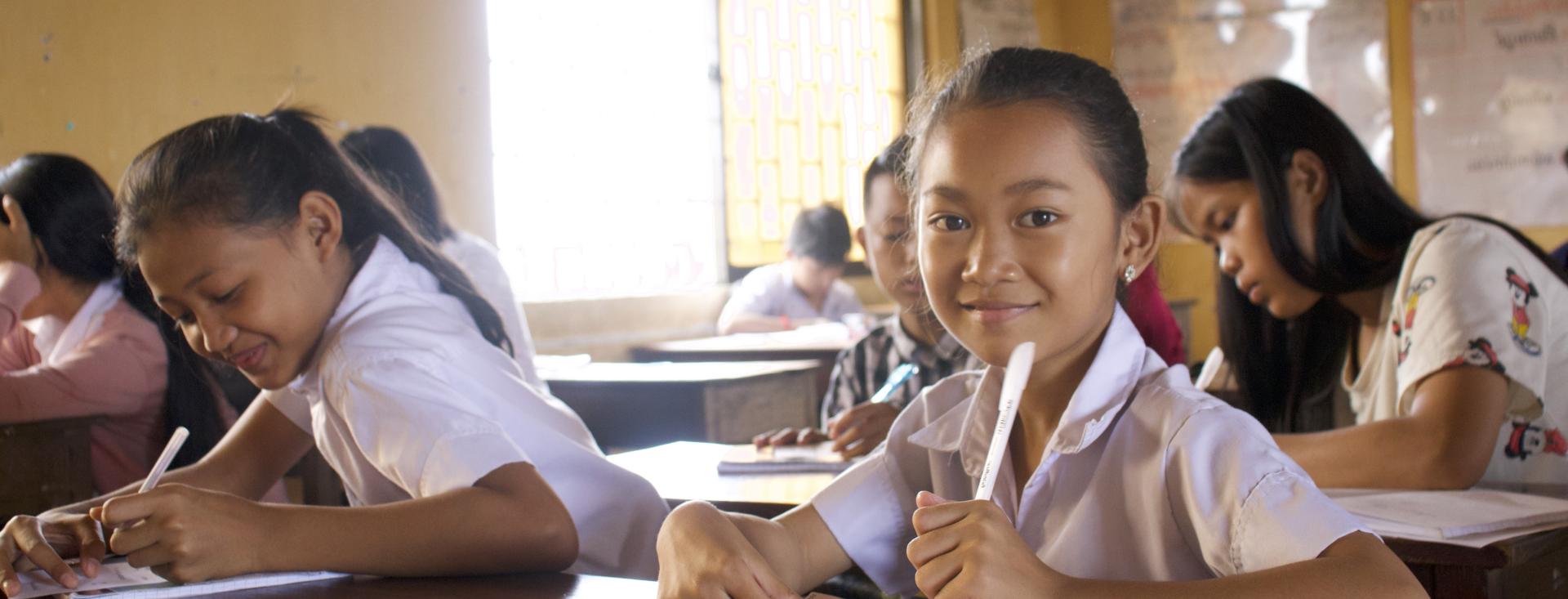 A young female Asian student is seated at a desk in a classroom, smiling as she looks directly into the camera