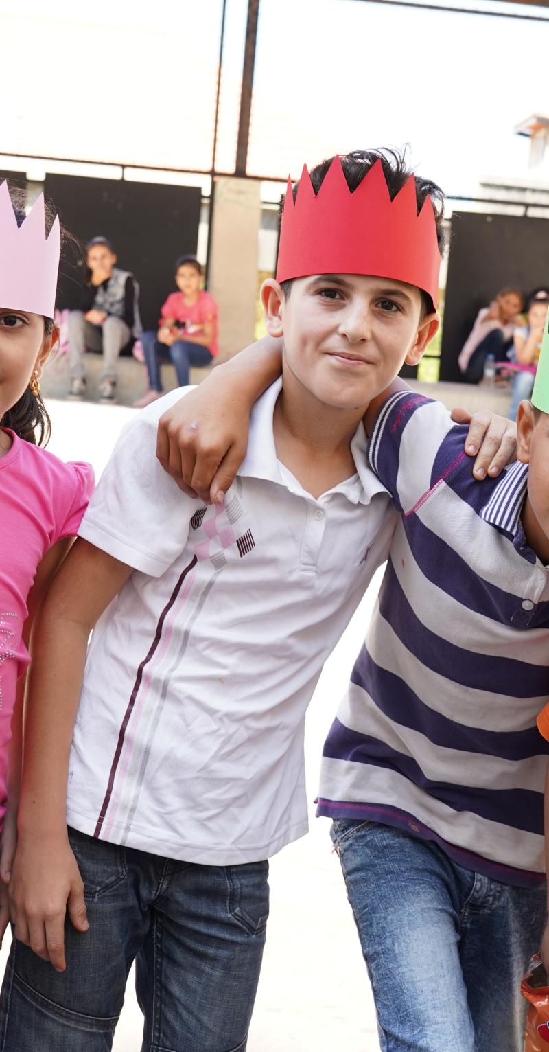 A group of 7 children wearing multi-colored paper crowns stand with their arms around each other's shoulders looking into the camera