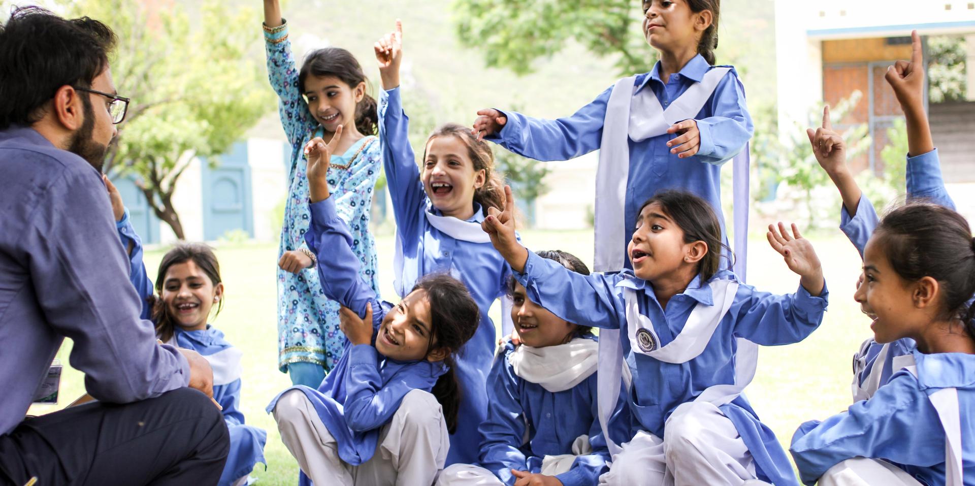 A group of girls in blue tunics with white sashes smile broadly with their hands in the air. Some are seated and some are standing.