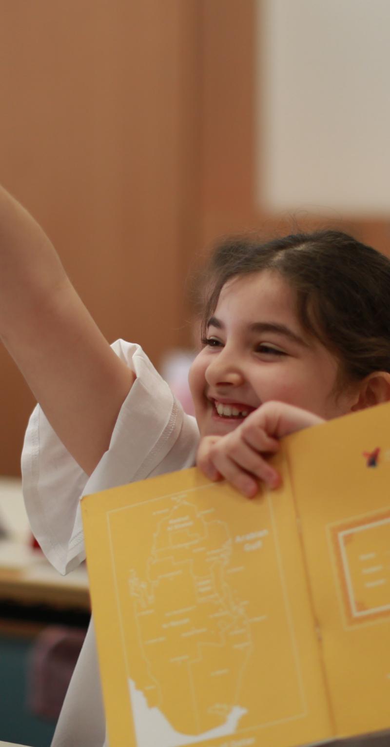 A smiling girl holidng an open book raises her hand excitedly