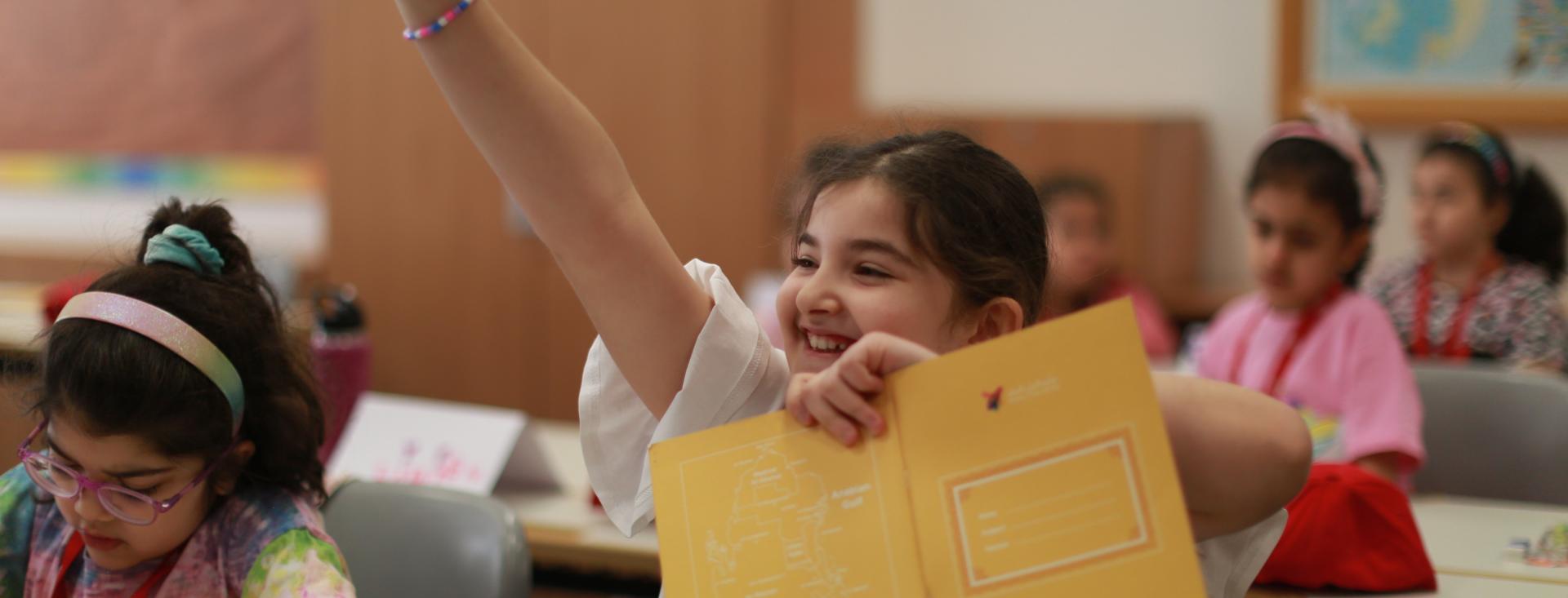 A smiling girl holidng an open book raises her hand excitedly