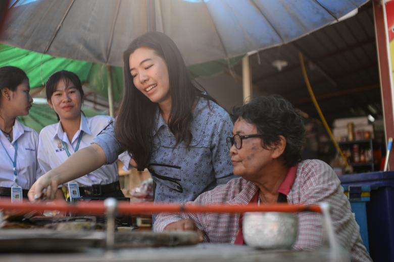 An elderly woman sits at a table while a young woman points something out to her and two students in uniform look on