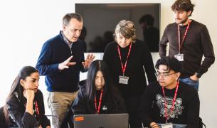 Three adults wearing name tags stand behind three teenage students, also wearing name tags, who surround a laptop 