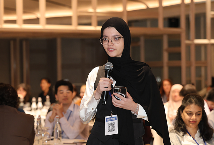 A young woman wearing glasses and a black Hijab speaks into a microphone as tables of adults behind her listen