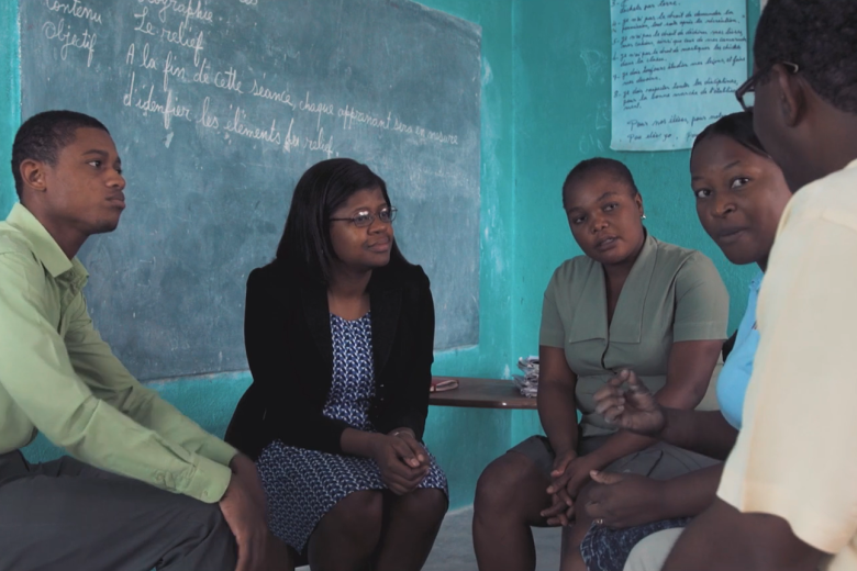 Three Black women and two Black men are seated in a semi-circle near a chalboard engaged in conversation