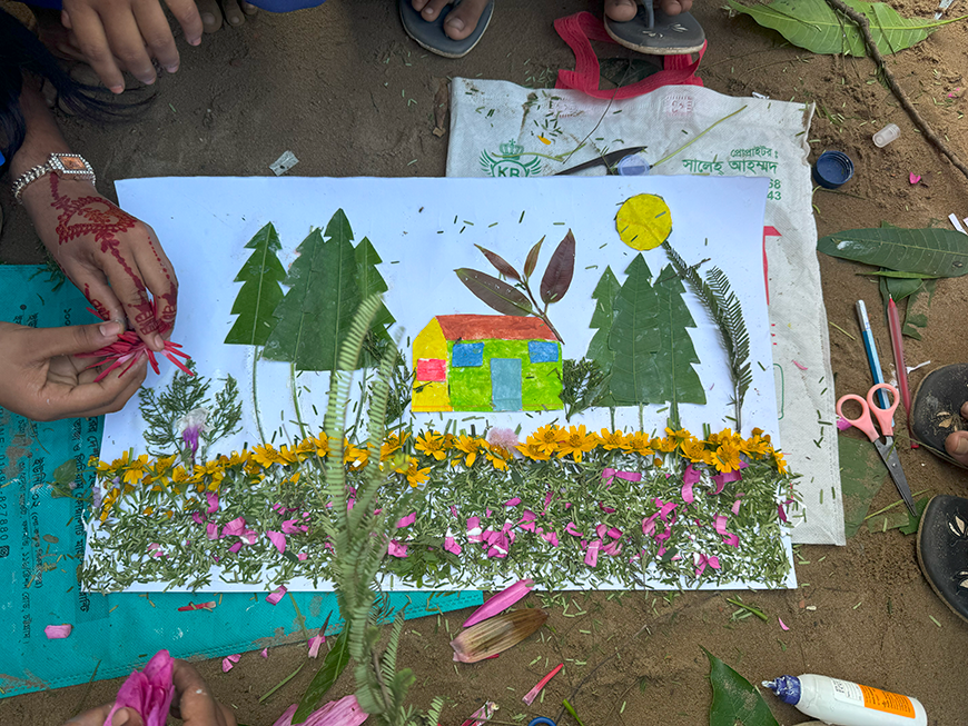 Children's hands and feet crowd around an artwork made of leaves and flowers depicting a house surrounded by trees and grass