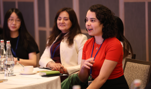 A young woman with shoulder-length brown curly hair and pale skin wearing a red shirt speaks while seated as others at the table watch and listen